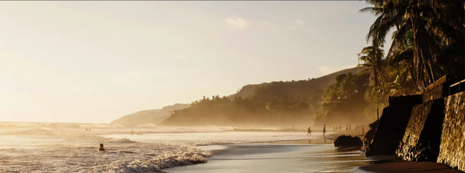 Waves rolling onto a sandy beach at sunset, with palm trees and hills behind